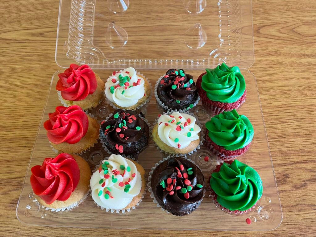 Open package of 12 mini cupcakes, some with green frosting, some with red, some with white or chocolate frosting with colored sprinkles.