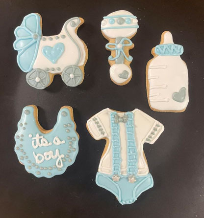 picture of gluten-free cutout sugar cookies decorated for a baby boy shower event.  Cookies decorated as a baby carriage, rattle, baby bottle, a bib and a onesie, all in baby blue and white frosting
