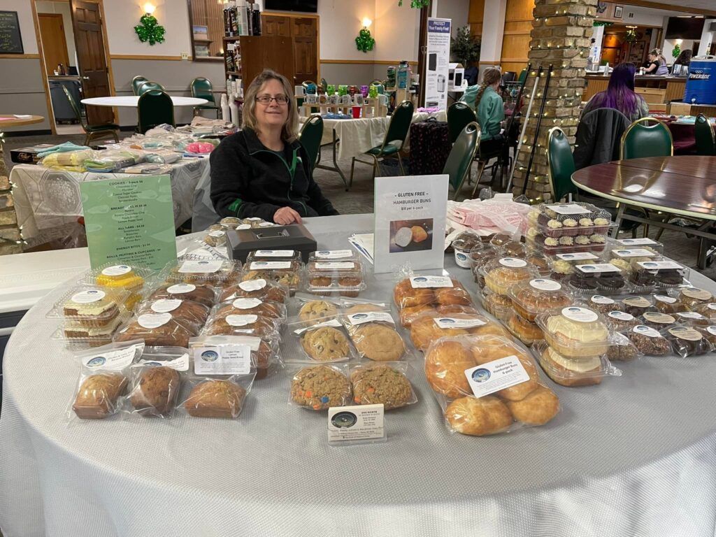 Mary seated behind a round table with gluten-free products laid out for sale in front of her